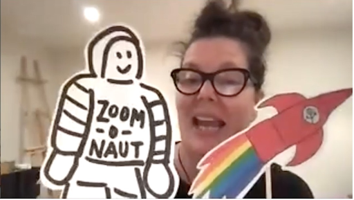 A person in glasses with long hair in a top bun appears to be speaking and is holding up an illustrated cut-out of a red rocket on a stick with rainbow flames coming out of it as well as a cut-out drawing of a smiling astronaut with the word ZOOM-O-NAUT written on its chest.