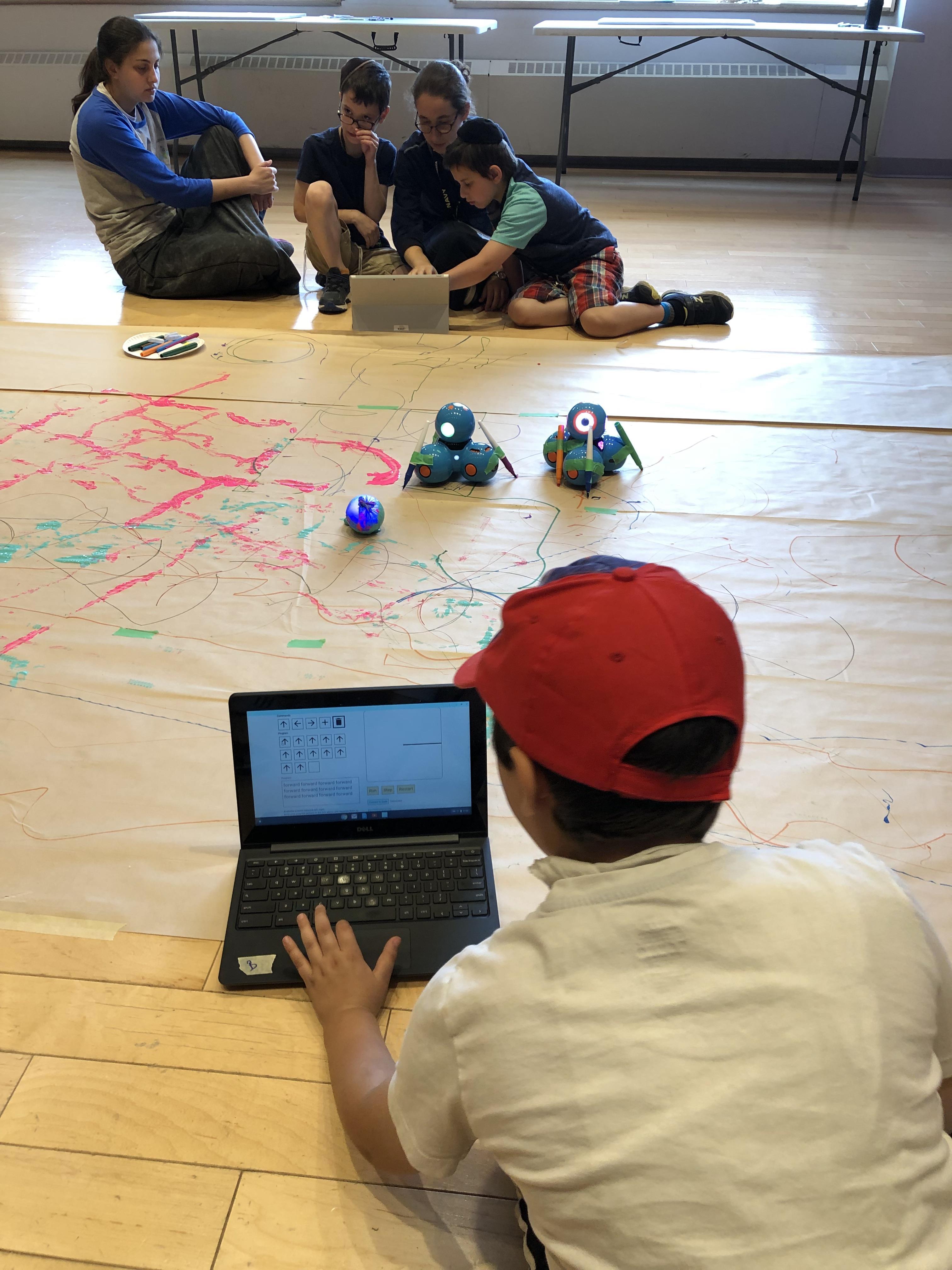 Facilitators and campers are working with the C2LC prototype on their laptops. They are creating programs for Dash and Sphero robots to draw shapes on large panels of paper taped on the floor. 