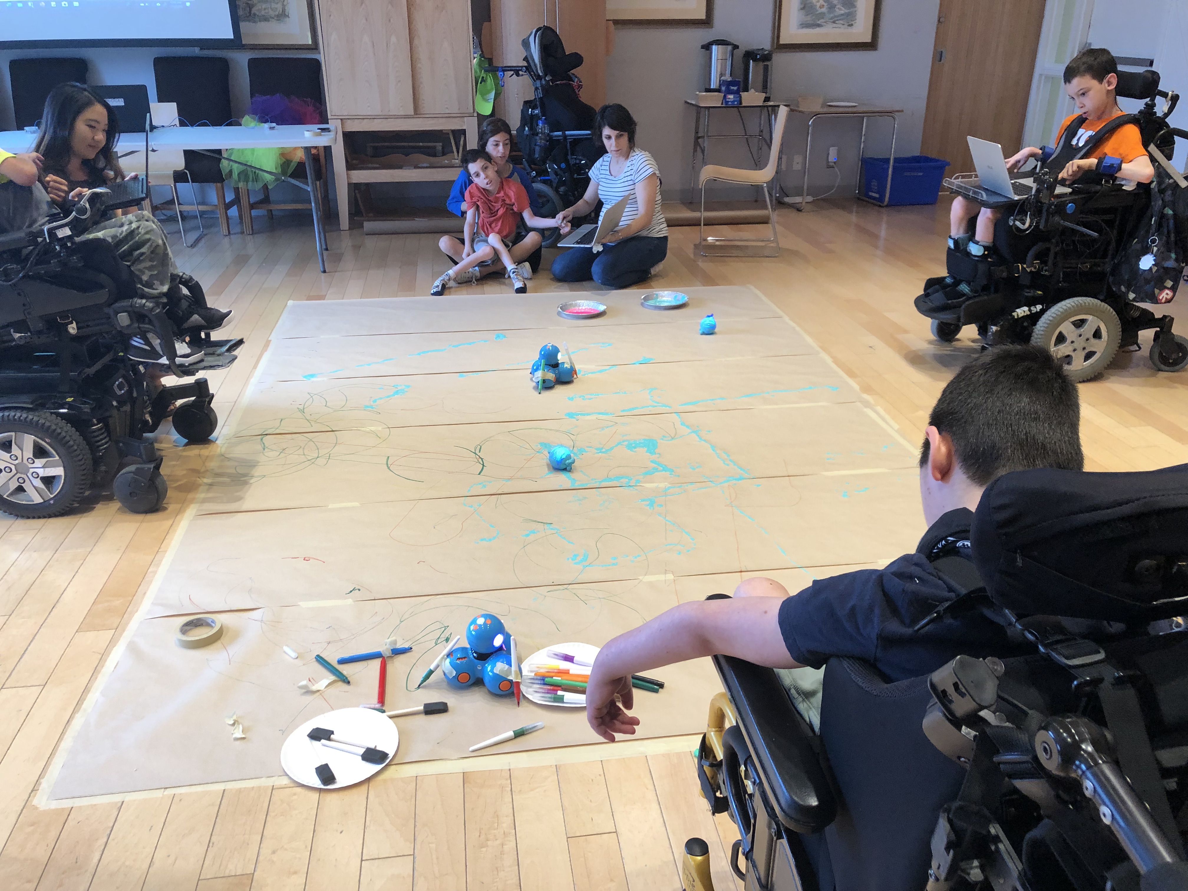 Campers sitting on the floor and using C2LC prototype on their laptops to code Dash and Sphero robots. Sphero is dipped in paint and markers are tapped to Dash, so they can draw shapes as they execute campers’ programs. 